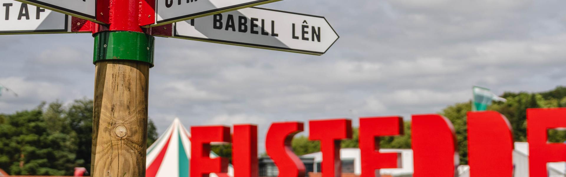 Eisteddfod 2024 sign in red under a cloudy sky with a fingerpost in the foreground on a grassy surface