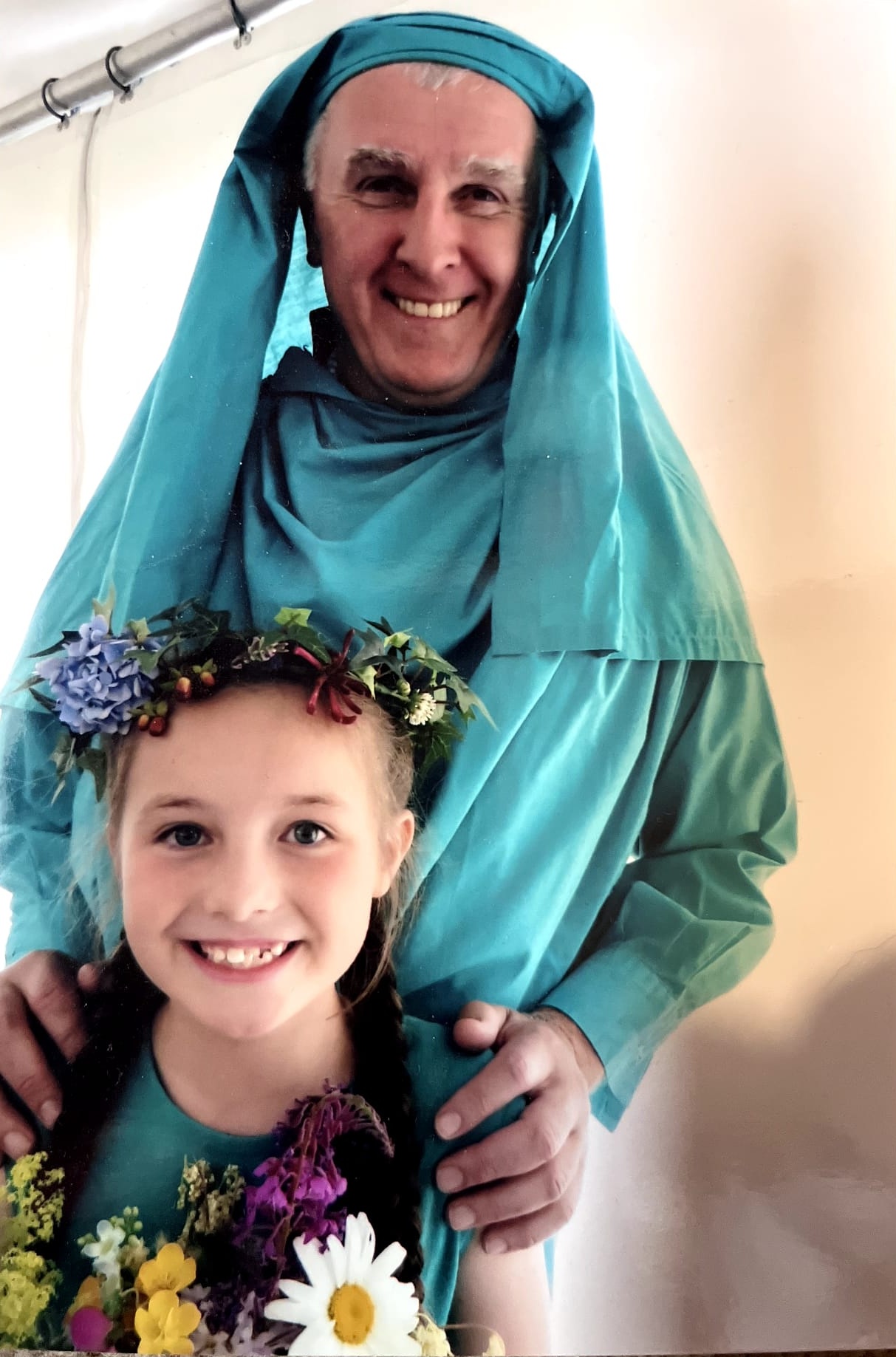 Two people standing together; one in the background wearing green Gorsedd robe and head-dress, with hands resting on the shoulders of the person in front, who is wearing a teal dress and a colourful flower crown