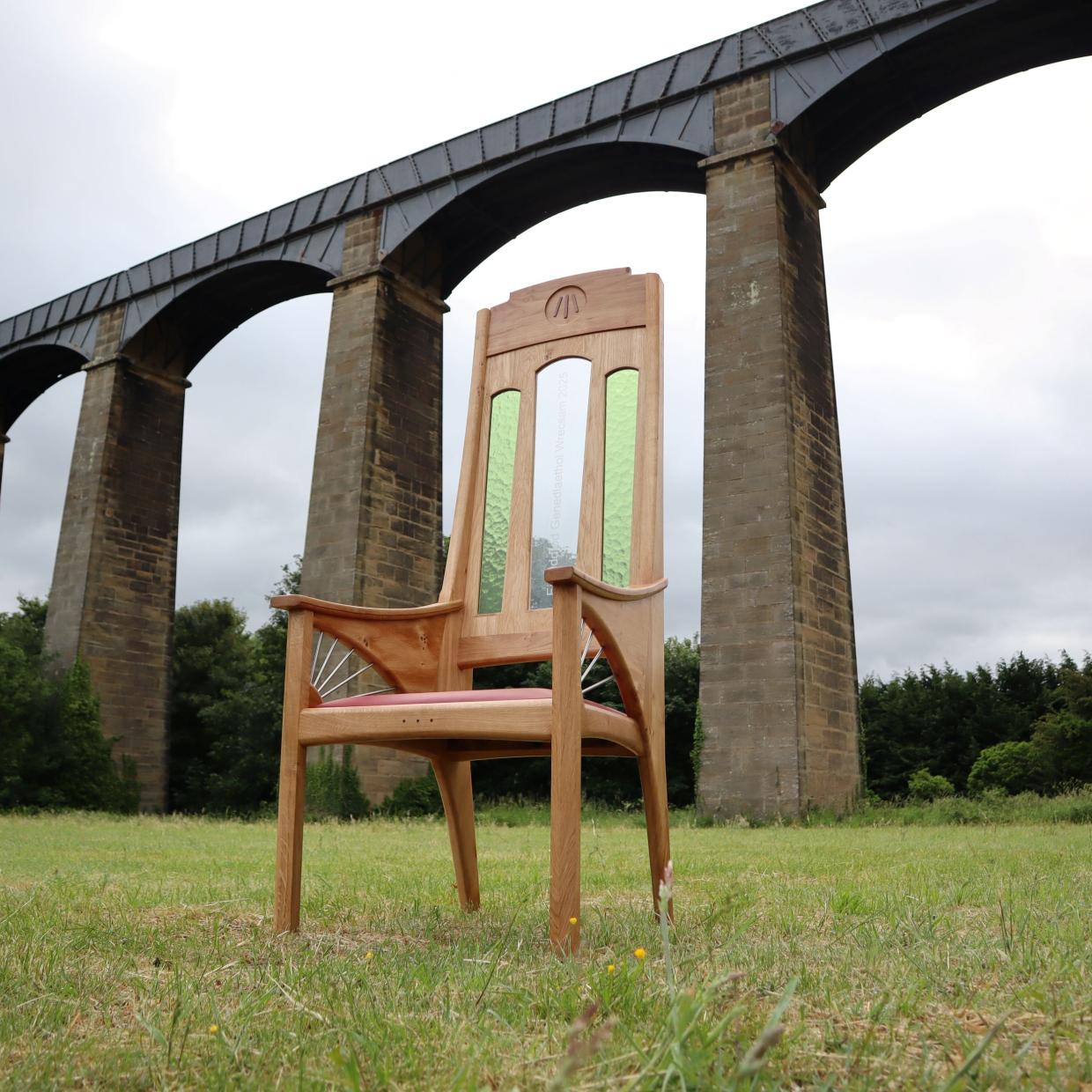 A wooden chair with green stained glass panels in the backrest sits on a grassy field in front of a large stone viaduct with multiple arches. The sky is overcast, and trees are visible in the background.