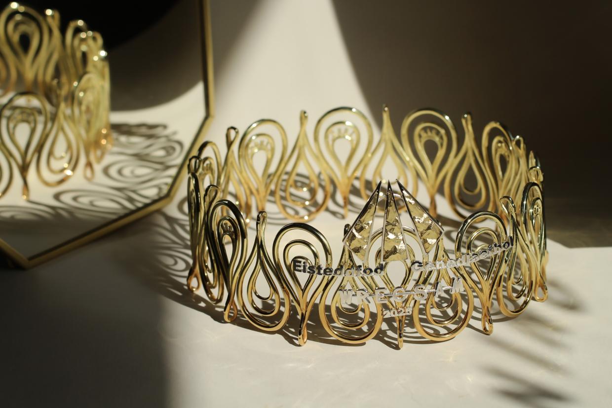 A golden crown with intricate, looping designs is placed on a surface with its reflection visible in a mirror behind it. The crown has text engraved on it that reads 'Eisteddfod Genedlaethol Cymru 2025'