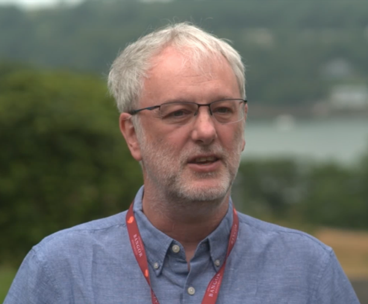 A person wearing glasses, a blue button-up shirt, and a red lanyard standing outdoors with greenery and water in the background
