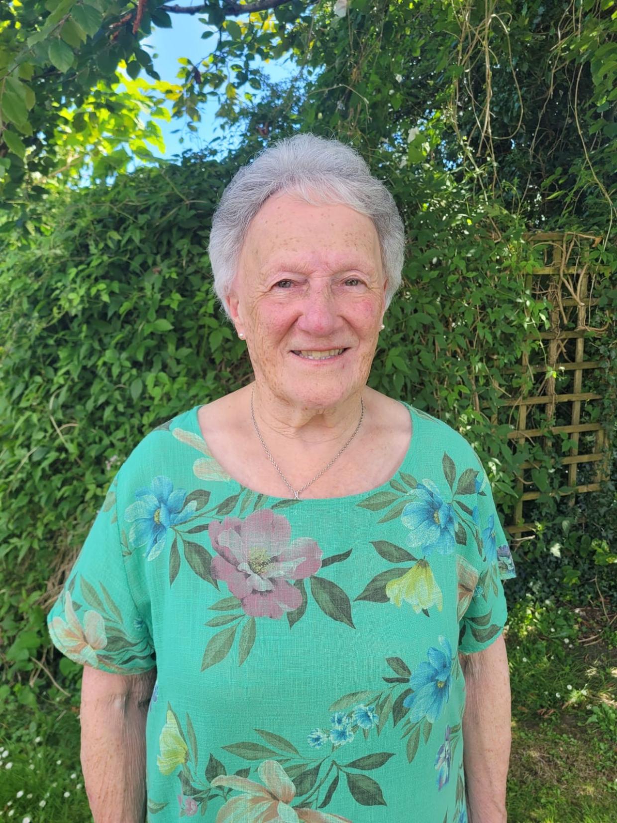 Person wearing a green floral shirt, standing in front of a lush green background with foliage and a wooden trellis.