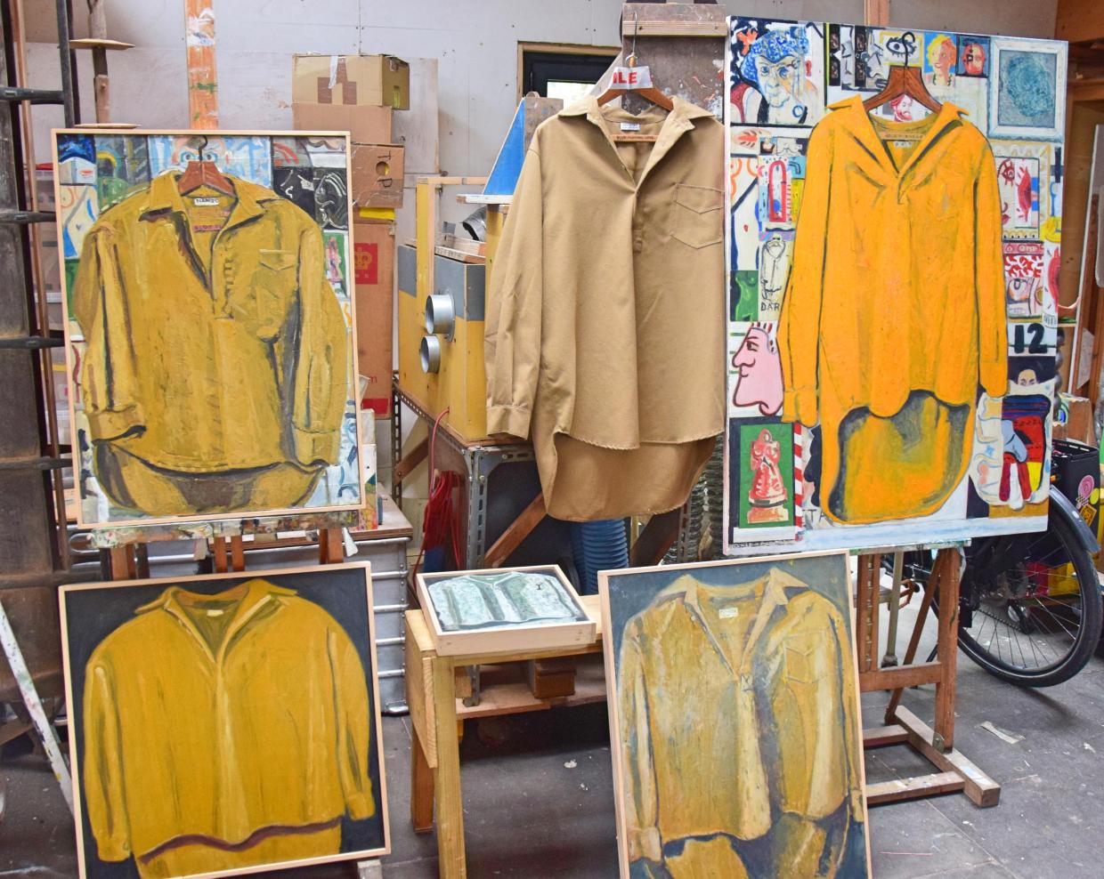 Art studio with multiple paintings of yellow shirts and a real beige shirt hanging on a hanger