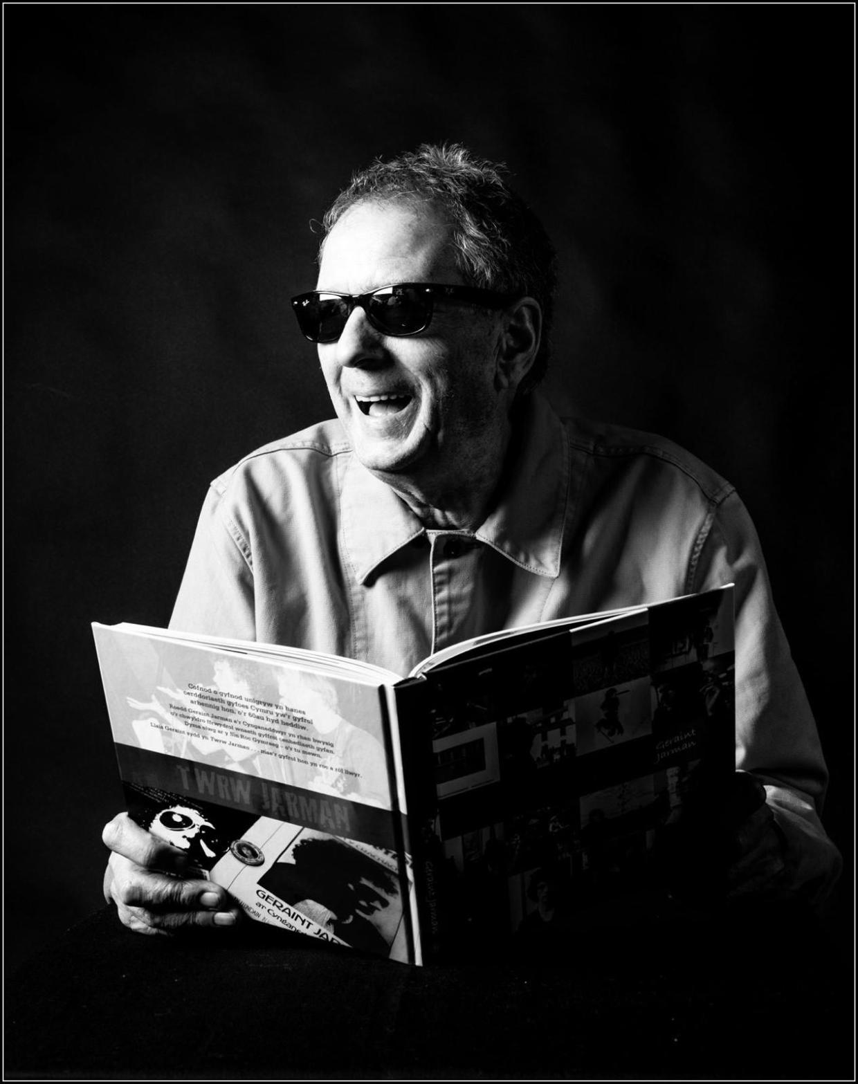 Black and white image of a person sitting and reading a book. The book cover features various images, including a photograph of someone wearing sunglasses