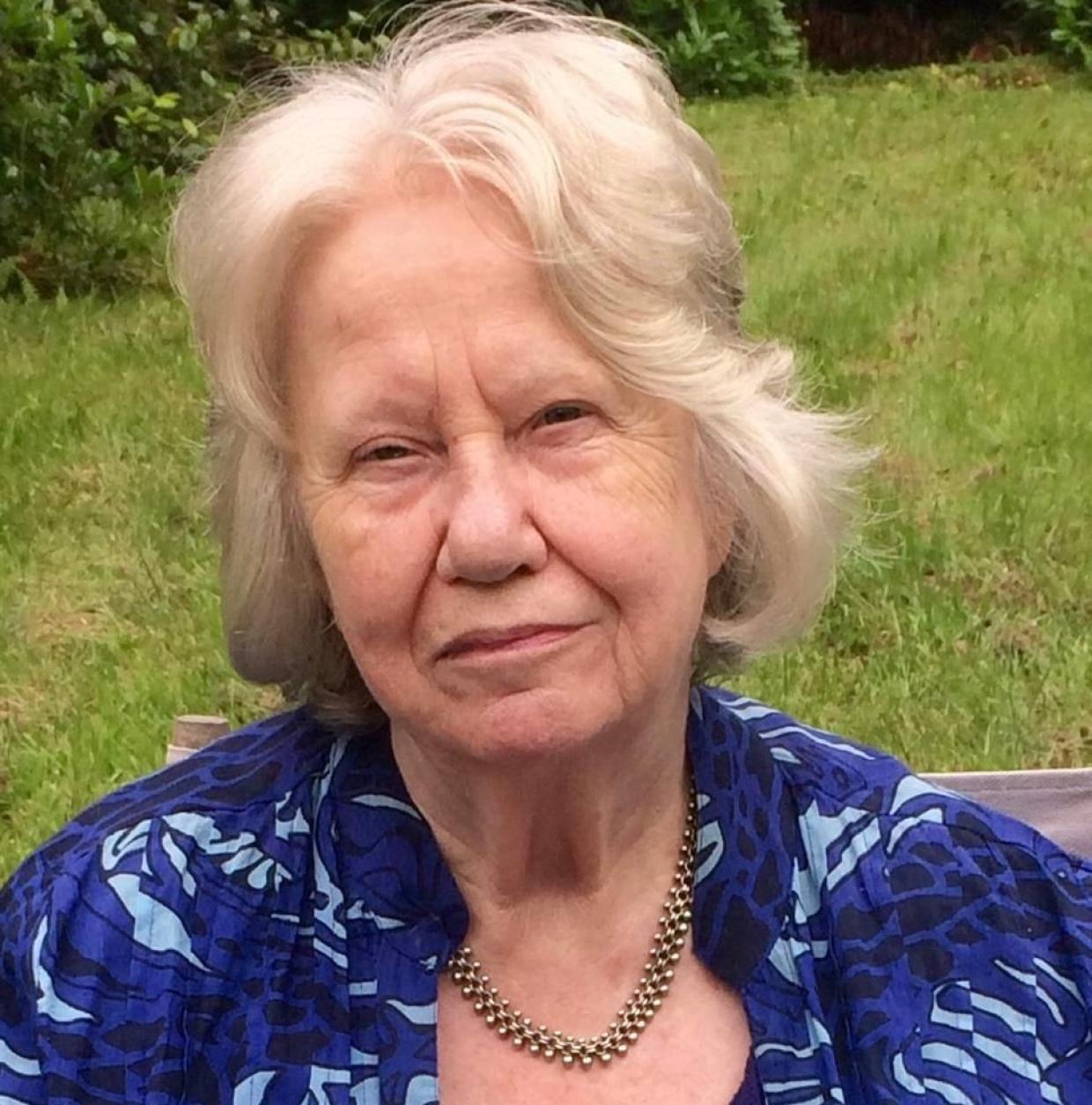 Woman with white hair wearing a blue patterned shirt and gold necklace, seated outdoors on grass
