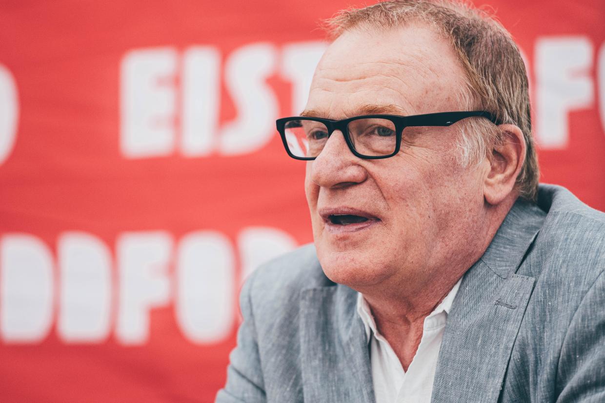 Person in a gray suit and glasses seated in front of a red background with partially visible white text reading Eisteddfod