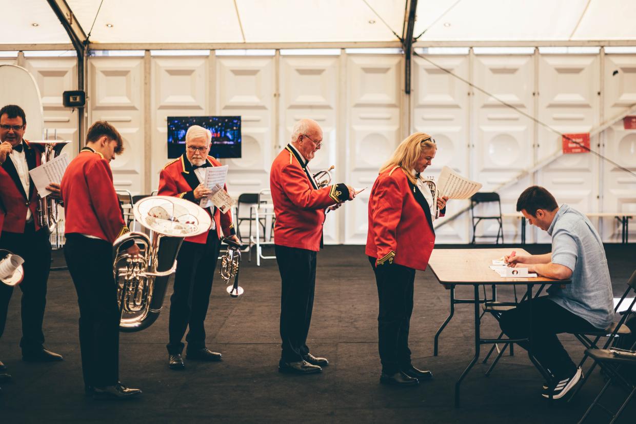 Group of people in red jackets holding musical instruments and sheet music, standing in line indoors; one person interacts with another seated at a table, possibly for event registration