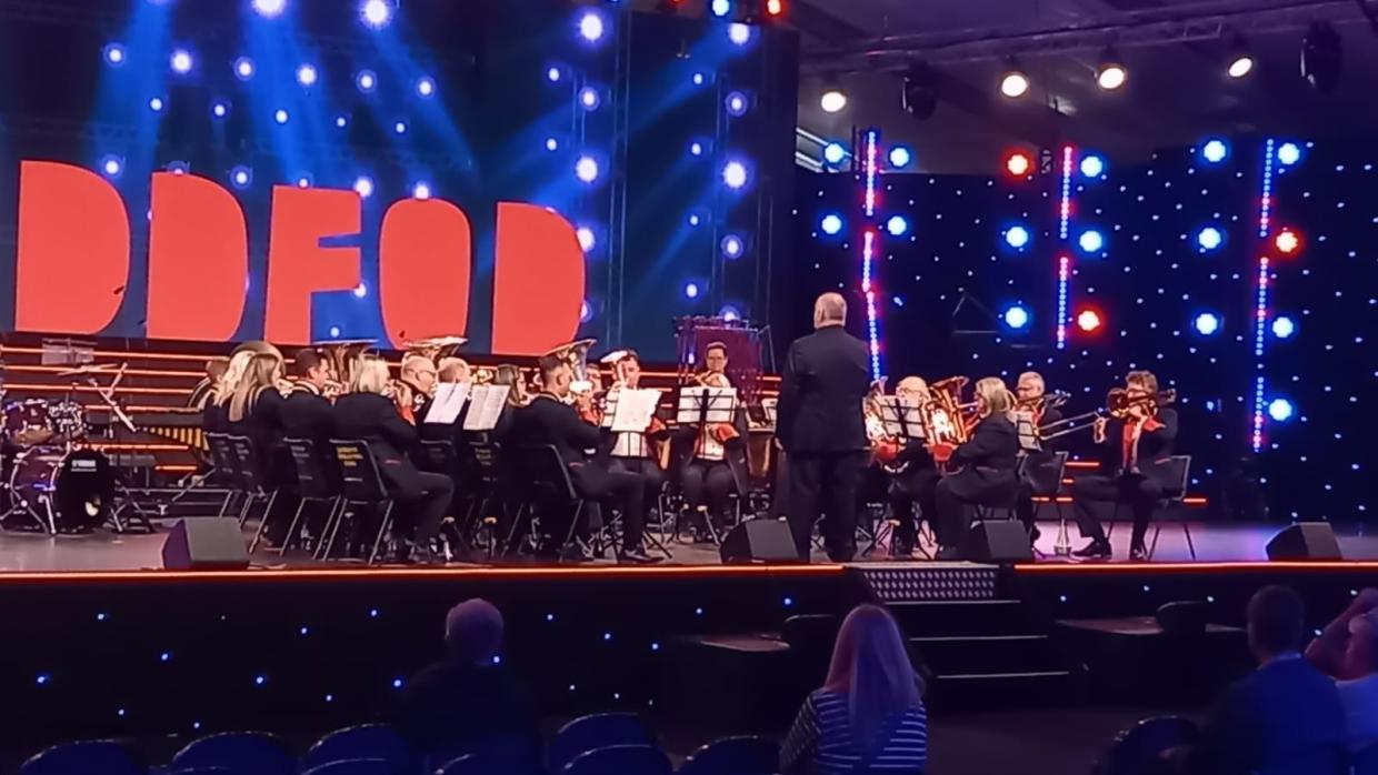 Brass band performing on stage with conductor leading; musicians seated in rows playing brass instruments, with large red 'EISTEDDFOD' letters and stage lights in the background