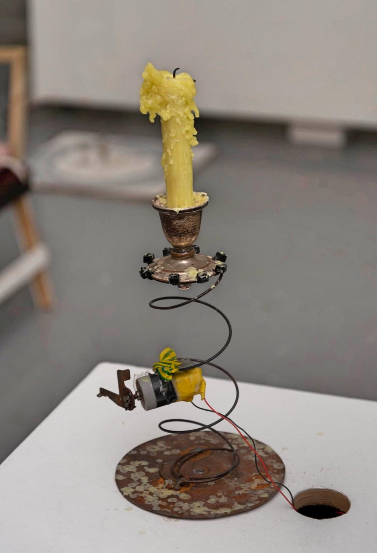 Yellow candle with melted wax on a metal holder, connected to a coiled wire spring and a small motor wrapped in yellow tape, mounted on a rusted circular metal base with extending wires