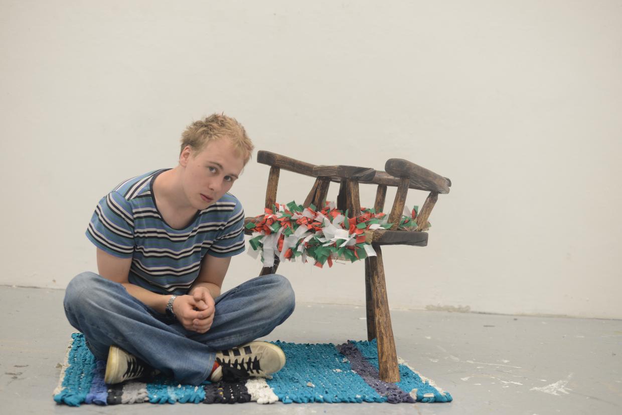 Person sitting cross-legged on a blue and black woven rug, wearing a striped shirt and jeans, next to a three-legged wooden chair decorated with red, white, and green fabric strips, against a plain white background.