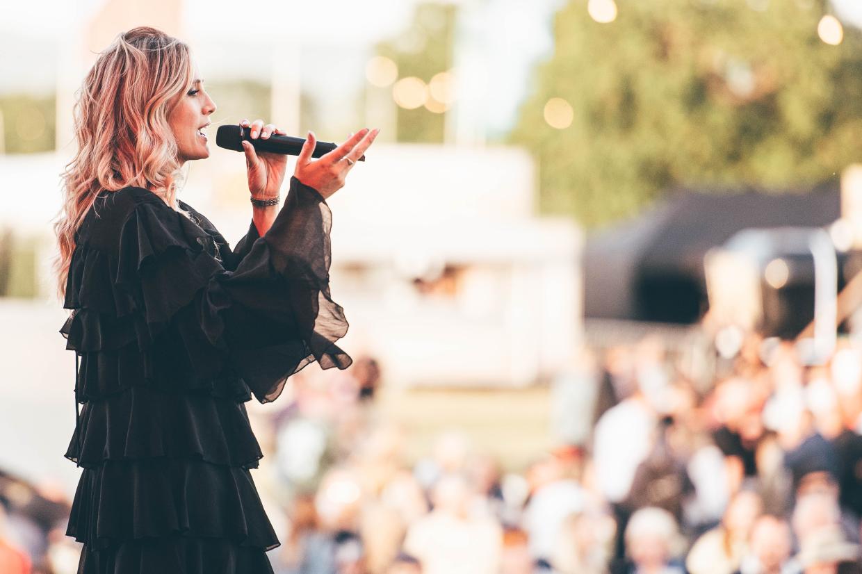 Person with long hair holding a microphone, speaking to an audience at an outdoor event, with face obscured and trees in the background
