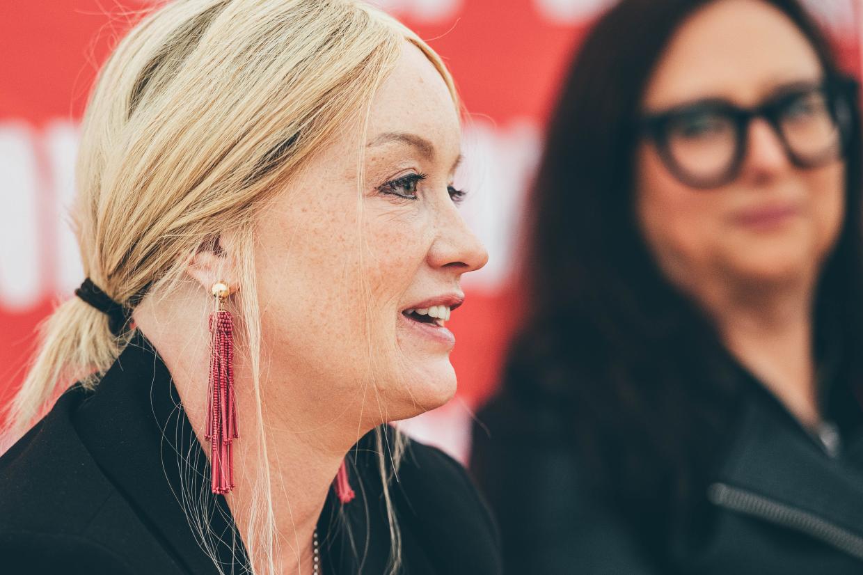 Two individuals; one with blonde hair and red tassel earrings, the other with dark hair in a black outfit, standing against a red background with partially visible white text