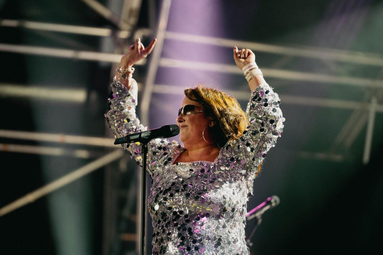 Person in sparkly outfit on stage with arms raised, microphone in front, and stage lighting and scaffolding in the background