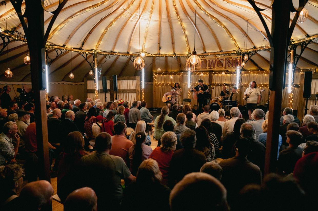 Audience seated inside a dome-shaped tent with string lights, watching musicians perform on a stage with guitars and microphones; TY GWERIN sign in background