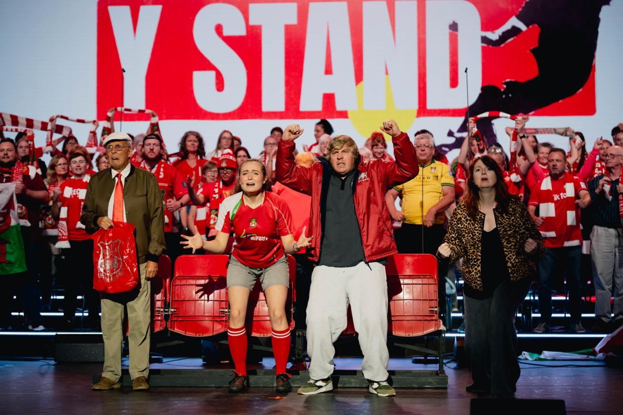 Group of sports fans in red and white cheering in front of a large 'Y STAND' sign, with four prominent individuals including an older man with a red bag, a person in sports gear, a casually dressed person with raised arms, and a woman in a leopard print jacket
