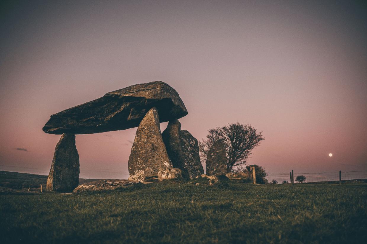 Twilight scene of a prehistoric dolmen with large upright stones supporting a flat capstone, set against a pink sky with the moon, grassy terrain, and distant tree and fencing