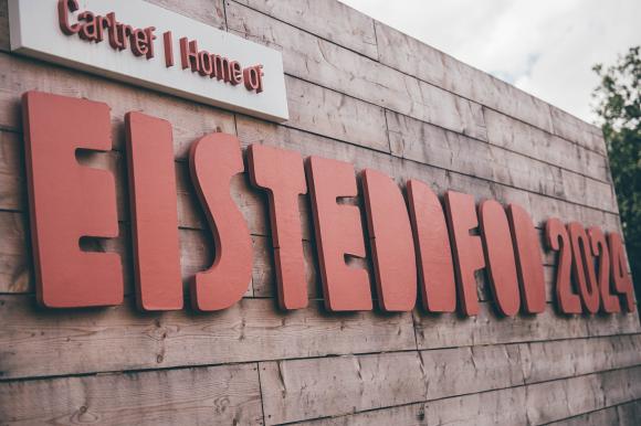 Eisteddfod 2024 sign in red on a wooden background under a cloudy sky
