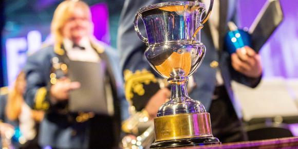 Silver trophy on display on a table on the stage with members of a brass band standing behind.  Purple background