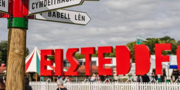 Eisteddfod 2024 sign in red under a cloudy sky with a fingerpost in the foreground on a grassy surface