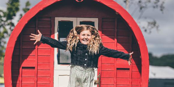 Young girl with long blonde hair jumping out of a red wooden caravan in a field with a cloudy sky