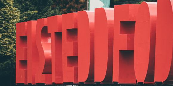 Large red 3D letters spelling 'EISTEDDFOD' displayed outdoors on a platform, with trees and a green and white flag in the background