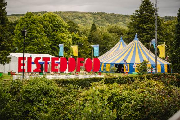 Colorful festival scene with a large blue and yellow striped tent and a red 'EISTEDDFOD' sign in front, surrounded by green trees, smaller tents, and flags under a cloudy sky
