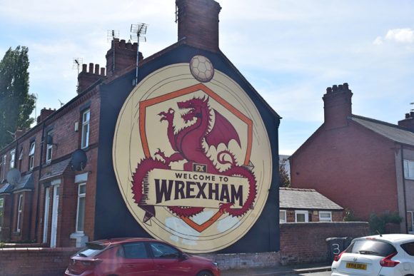 Large mural on the side of a brick house featuring a red dragon with wings spread inside an orange-bordered shield, with a soccer ball above and the words 'WELCOME TO WREXHAM' below. Houses and two parked cars are visible in the background.