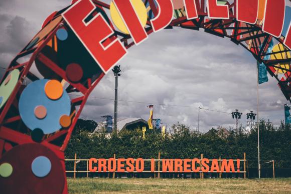 Colorful arch with 'EISTEDDFOD' written on it, and a sign below reading 'CROESO I WRECSAM!' against a backdrop of greenery and cloudy skies