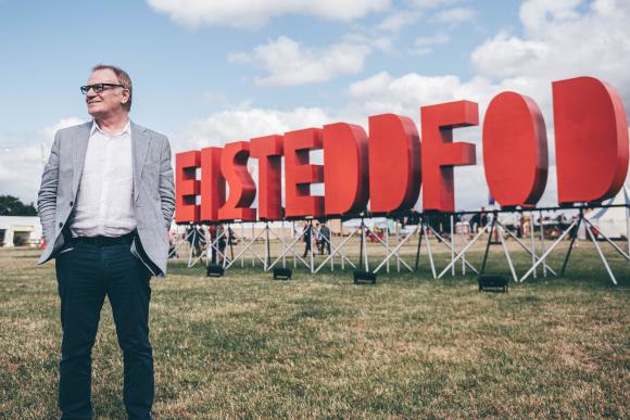 Man in grey blazer, white shirt, and dark pants standing on grass in front of a large red 'EISTEDDFOD' sign with a partly cloudy sky and tents in the background