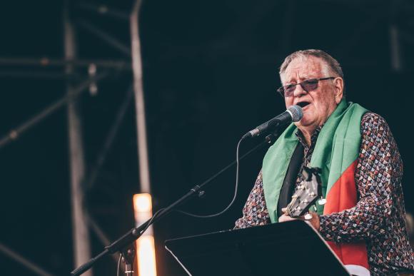 Person on stage holding a guitar and singing into a microphone, wearing a patterned shirt and draped in a Palestinian flag, with stage equipment and lighting in the background