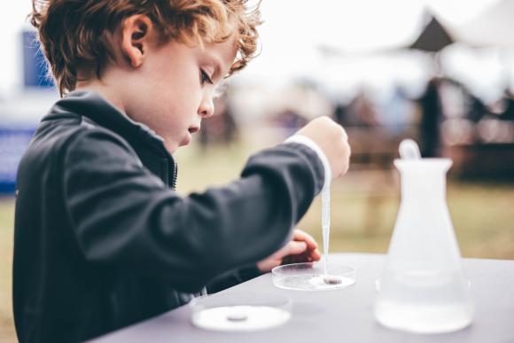 Child in dark jacket using a dropper to place liquid into a petri dish, with a conical flask nearby, in an outdoor science activity setting