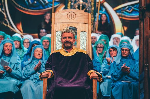 Person in a dark purple and gold robe seated on an ornate wooden throne, with individuals in blue robes behind them, set against a richly decorated background.