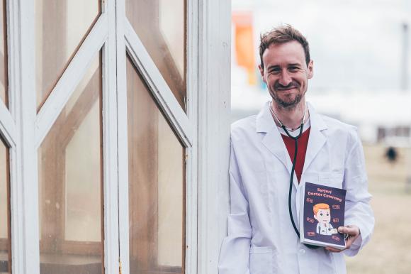 Person in a white lab coat holding a prescription pad titled 'Syrjeri Doctor Cymraeg',and a building with glass windows in the background