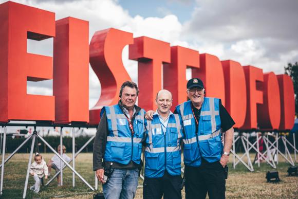 Three volunteers in blue reflective vests standing in front of large red 'EISTEDDFOD' letters, with children playing on the grass in the background under a cloudy sky