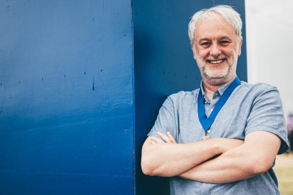 Person with crossed arms wearing a gray shirt and blue lanyard, standing next to a blue wall.