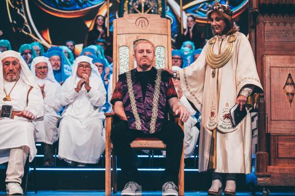 Ceremonial scene at the Eisteddfod with a person seated on an ornate wooden Chair, surrounded by individuals in white robes and a figure in ceremonial attire holding a book