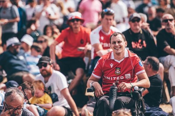Group of people gathered outdoors, with a person in a red sports jersey seated in a wheelchair in the foreground, and others standing or sitting in casual clothing