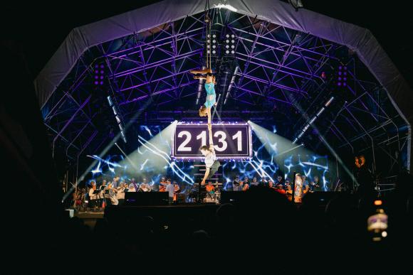 Stage performance with acrobats suspended in the air, large digital display showing '2131', musicians and performers on stage, under bright lights and metal framework