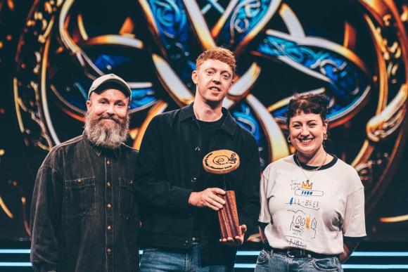 Three individuals standing in front of a colorful decorative background, with the central figure holding a wooden award or trophy;