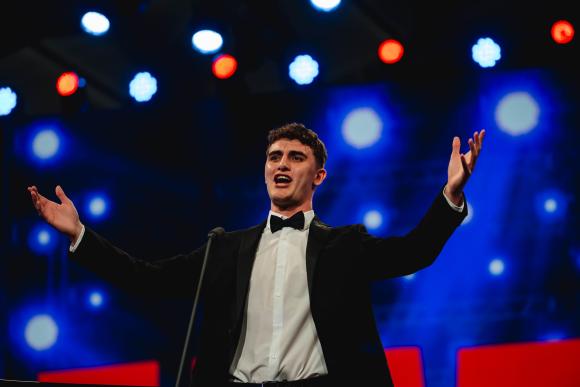 Person in formal black suit with bow tie standing with arms outstretched under bright stage lights and blue lighting effects