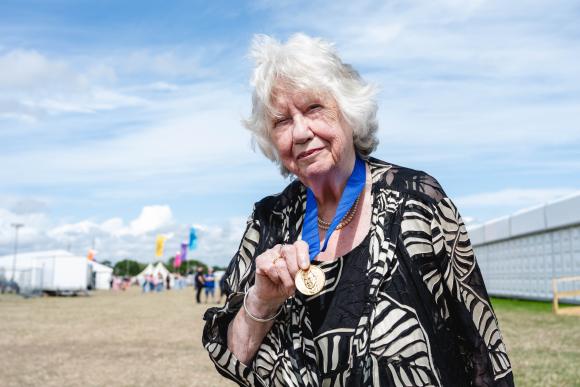 Person with white hair holding a gold medal, wearing a black and white patterned outfit with a blue ribbon, in an outdoor setting with tents and people in the background