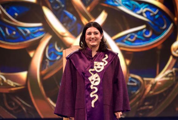 Person in a purple robe with theatrical masks and snake design, standing before an ornate, colourful patterned background.