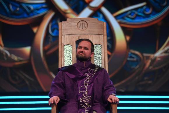 Person in a purple and gold embroidered robe seated on an intricately carved wooden chair, with a vibrant patterned background