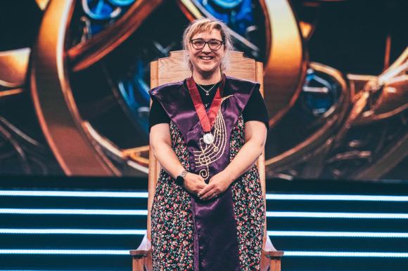 Person wearing a floral dress and purple robe with gold embroidery, seated on a wooden throne-like chair with a medal around their neck, against a decorative background with circular patterns and blue accents