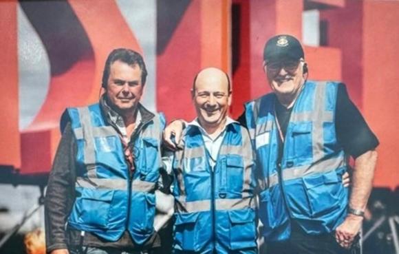 Three individuals in blue reflective safety vests standing side by side in front of large red and white structures