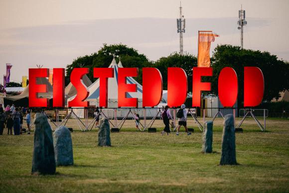 Outdoor event with large red 'EISTEDDFOD' letters, people walking among tents and trees, and a circle of standing stones in the foreground