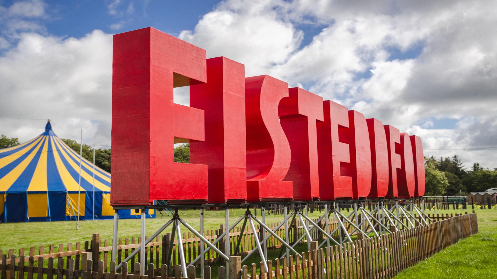 Arwydd Eisteddfod gyda phafiliwn streipiog melyn a glas yn y cefndir