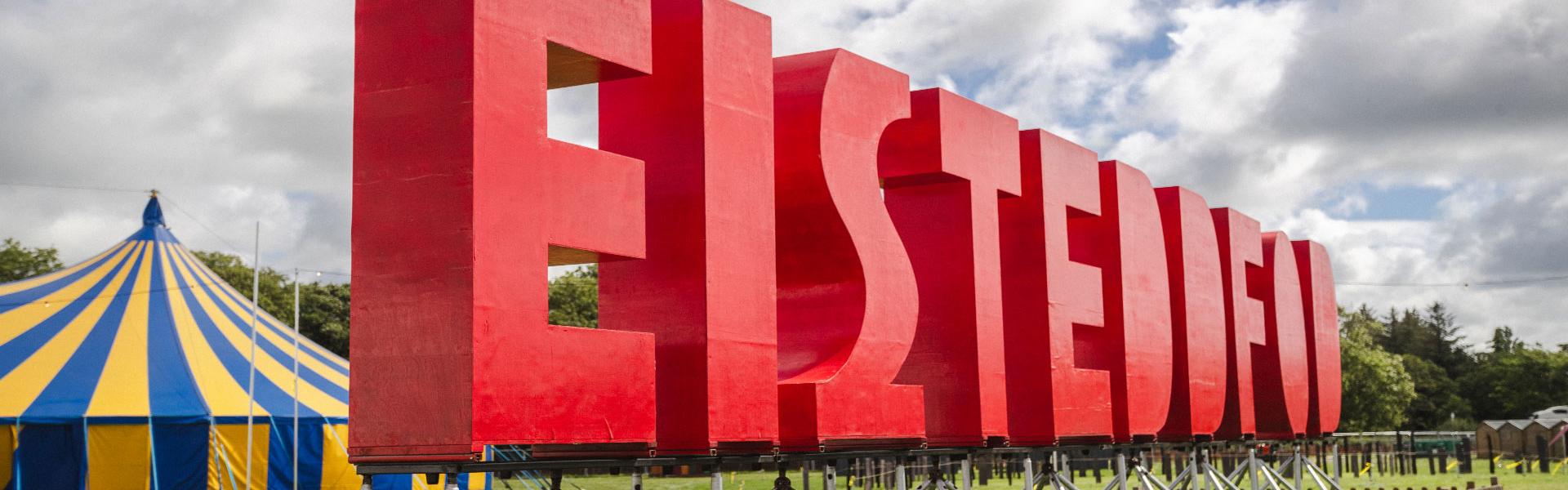 Huge red letters spelling out 'Eisteddfod' at an angle in a field with a striped pavilion in the background and a cloudy sky above