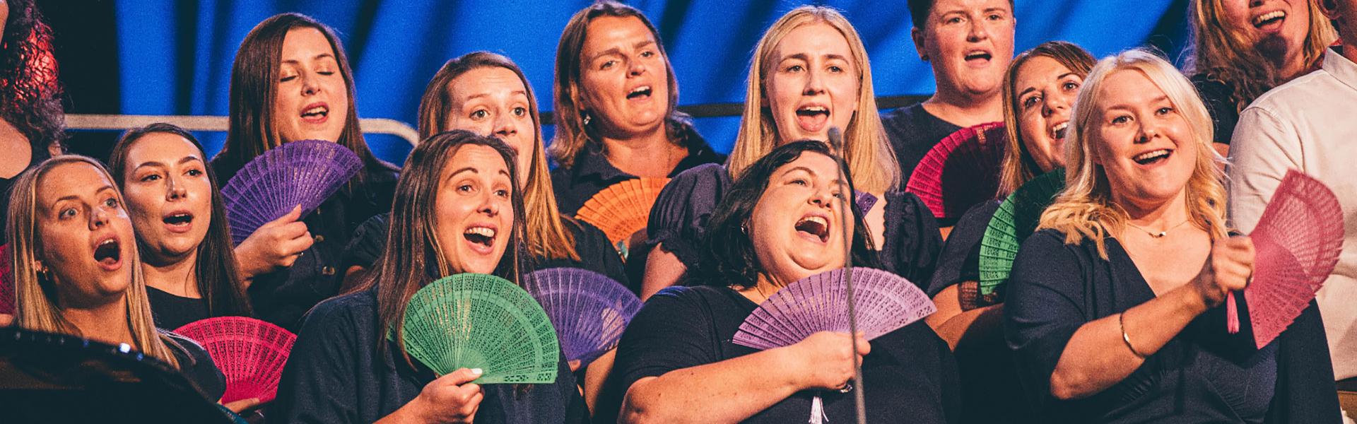 Choir performing on the pavilion stage with 'fans' in their hands and smiling