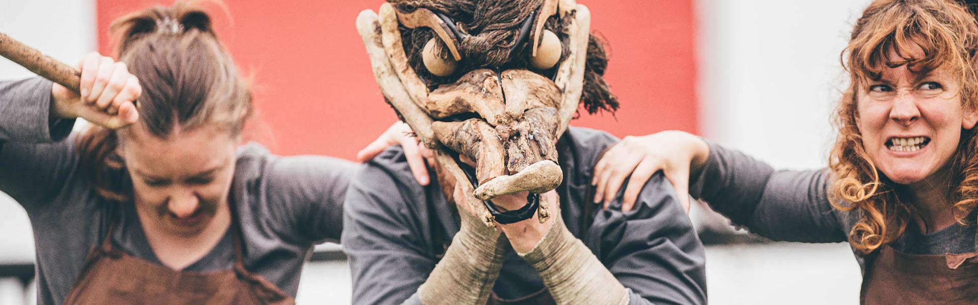 a street theatre show with the performers wearing scary animal masks in front of a red Eisteddfod sign